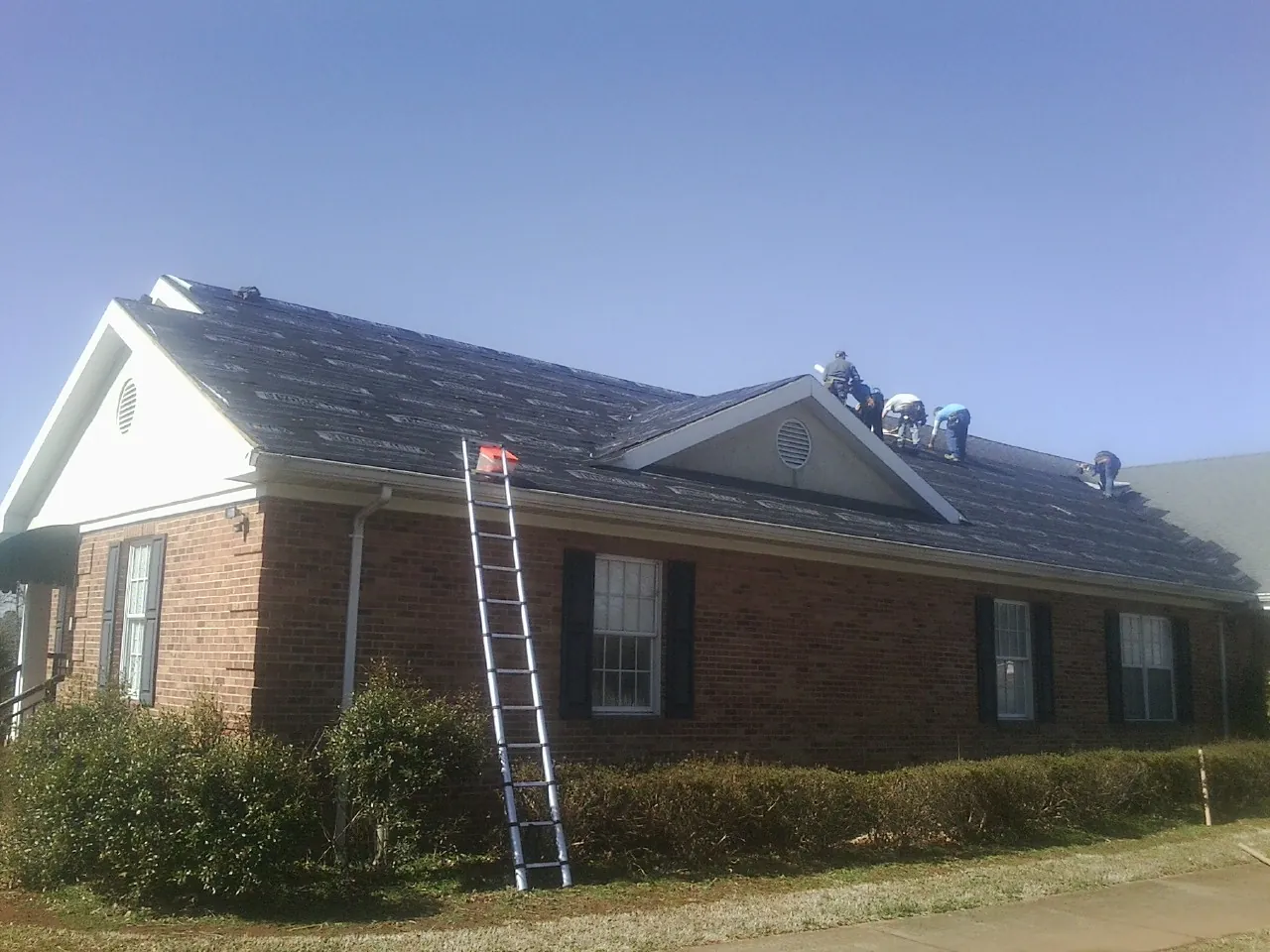 Crew tearing shingles off a brick ranch roof with a ladder set — Metal Roof Repair work by our crew in Manchaca