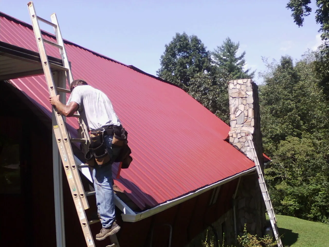 Roofer on a ladder beside a red metal roof and stone chimney — Soffit & Fascia Repair by Roofing Brothers in Manchaca